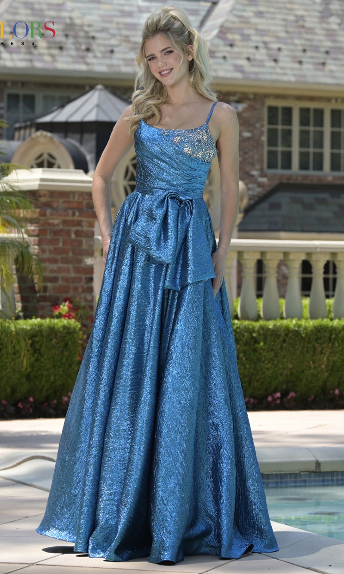 A woman stands by a pool wearing the Colors Dress 3793, a shimmering blue, sleeveless, floor-length A-line prom gown with a decorative bow and beaded bodice.